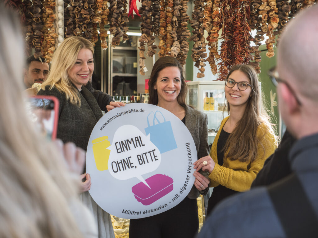 Foto von Stephanie Jacobs, Kristina Frank und Johanna Koch, die einen Riesensticker von „Einmal ohne, bitte“ in einem Lebensmittelgeschäft hochhalten